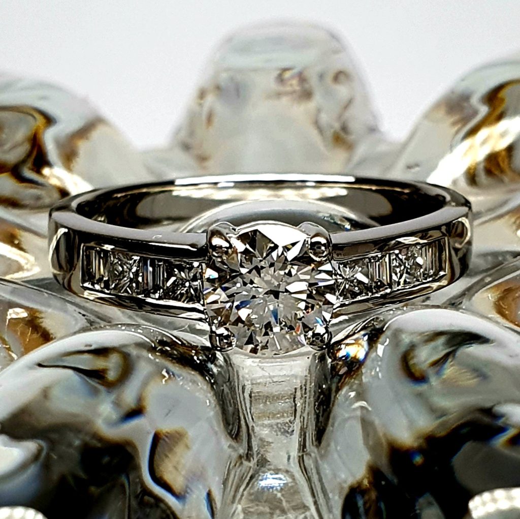 The Lambeth Platinum Diamond Engagement Ring RPT00045 The centre is set with a 0.70ct round brilliant-cut diamond of Colour H clarity SI1, which has an excellent cut, proportions, and symmetry. The side shoulders are channel set with a total of four princess-cut diamonds and four baguette-cut diamonds. The total diamond weight is 1.08 carats.