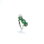 A captivating image of a Three Cluster Emerald Ring in 18ct white gold. The ring showcases three clusters of vibrant green emeralds, with six emeralds in each cluster. Adorning the ring shank is a graceful wave design, adding a touch of elegance to the overall aesthetic. The clusters are accentuated by three sparkling diamonds, positioned at the centre of each cluster." Angled top view.