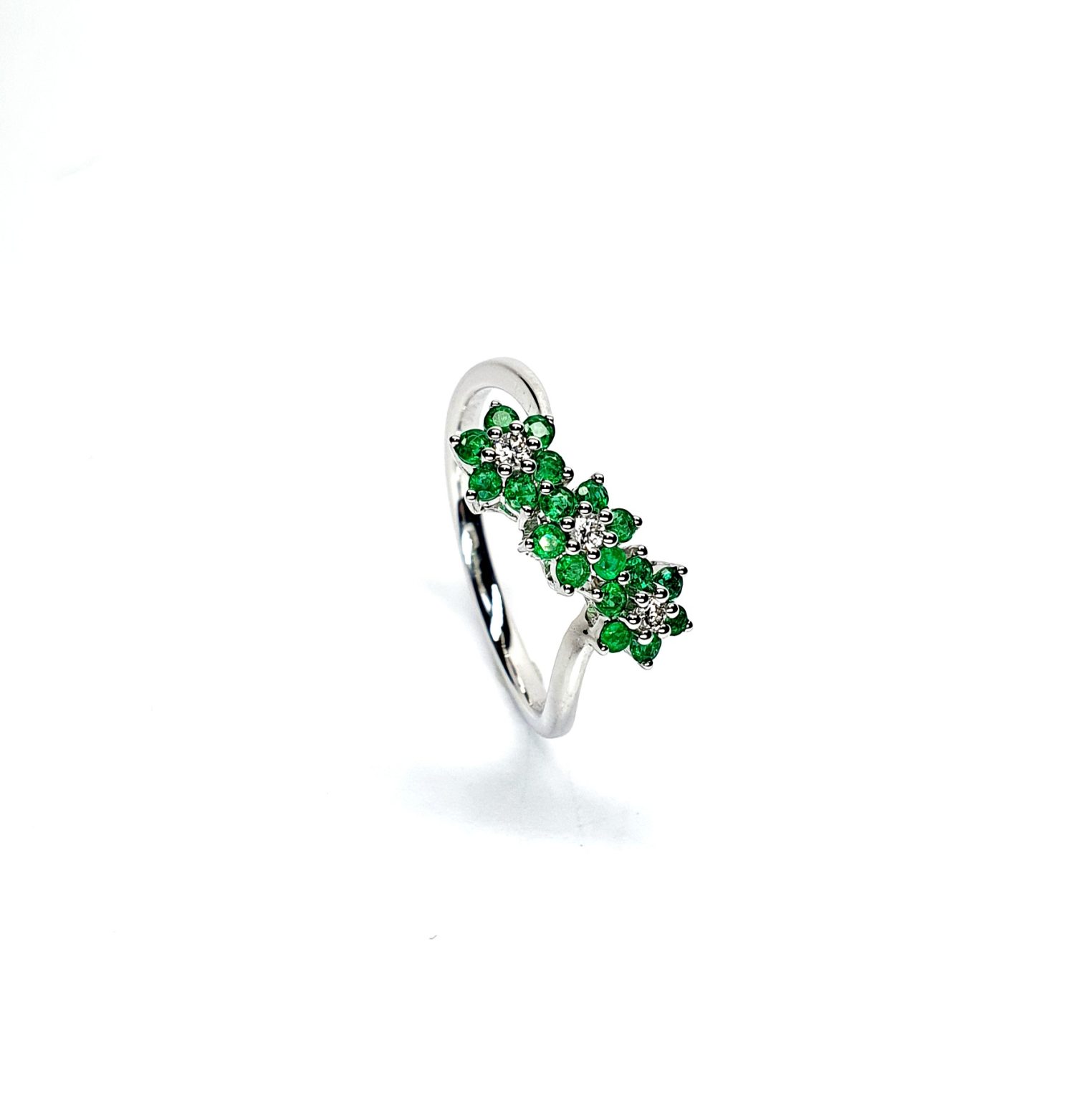 A captivating image of a Three Cluster Emerald Ring in 18ct white gold. The ring showcases three clusters of vibrant green emeralds, with six emeralds in each cluster. Adorning the ring shank is a graceful wave design, adding a touch of elegance to the overall aesthetic. The clusters are accentuated by three sparkling diamonds, positioned at the centre of each cluster." Angled top view.