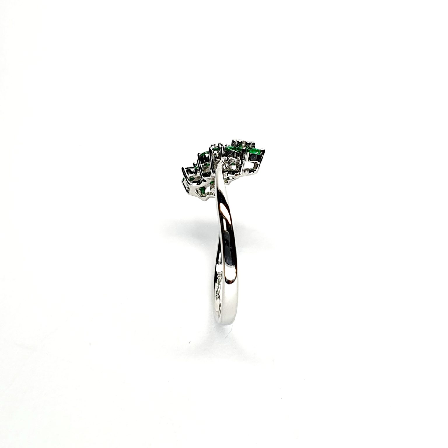 A captivating image of a Three Cluster Emerald Ring in 18ct white gold. The ring showcases three clusters of vibrant green emeralds, with six emeralds in each cluster. Adorning the ring shank is a graceful wave design, adding a touch of elegance to the overall aesthetic. The clusters are accentuated by three sparkling diamonds, positioned at the centre of each cluster." Edge view.