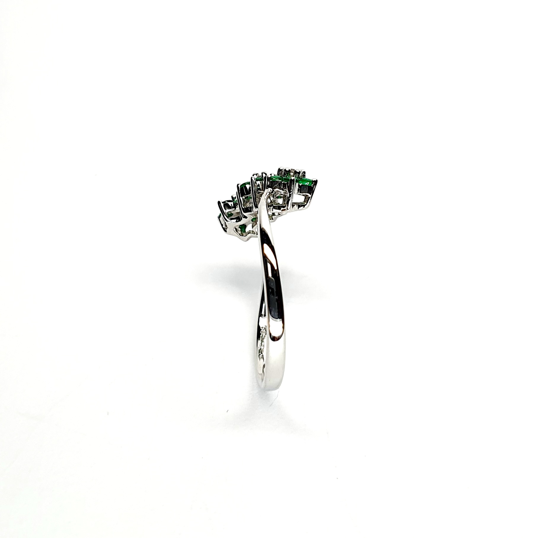 A captivating image of a Three Cluster Emerald Ring in 18ct white gold. The ring showcases three clusters of vibrant green emeralds, with six emeralds in each cluster. Adorning the ring shank is a graceful wave design, adding a touch of elegance to the overall aesthetic. The clusters are accentuated by three sparkling diamonds, positioned at the centre of each cluster." Edge view.