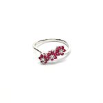 A captivating image of a Three Cluster Ruby Ring in 18ct white gold. The ring showcases three clusters of vibrant rubies, with six rubies in each cluster. Adorning the ring shank is a graceful wave design, adding a touch of elegance to the overall aesthetic. The clusters are accentuated by three sparkling diamonds, positioned at the centre of each cluster." Lying flat view.