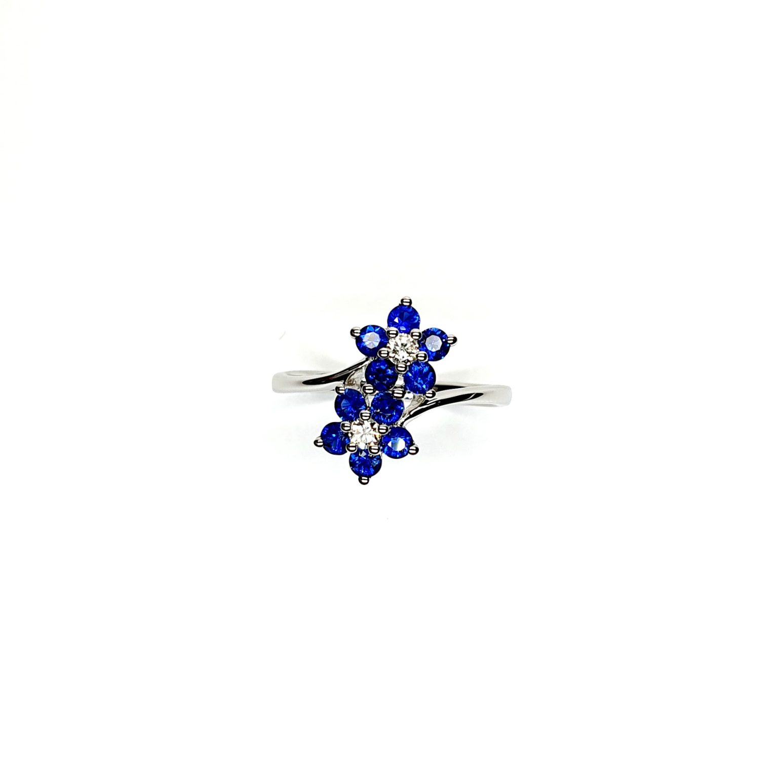 "An elegant 18ct white gold ring with a crossover shank featuring two clusters of five round bright blue sapphires, each cluster surrounds a single white sparkling diamond, creating a captivating and sophisticated design." Top View.