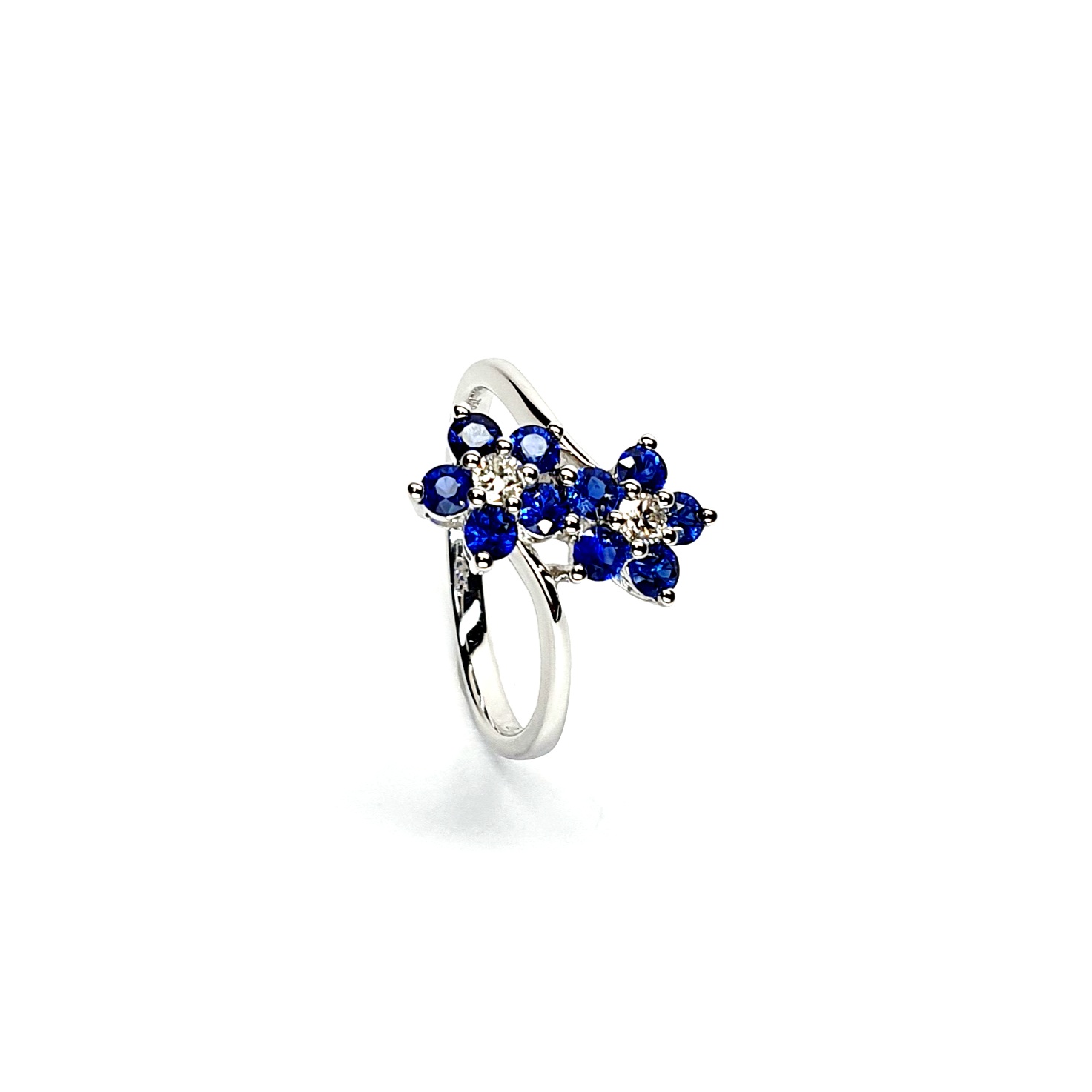"An elegant 18ct white gold ring with a crossover shank featuring two clusters of five round bright blue sapphires, each cluster surrounds a single white sparkling diamond, creating a captivating and sophisticated design." Angled View.