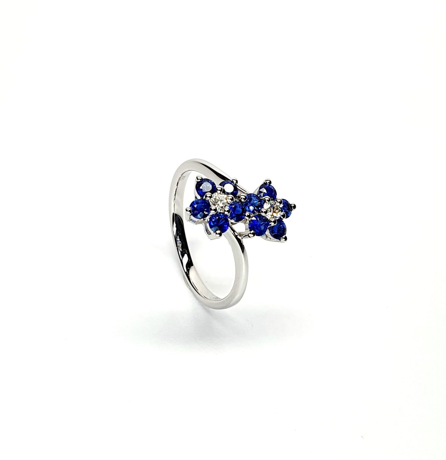 "An elegant 18ct white gold ring with a crossover shank featuring two clusters of five round bright blue sapphires, each cluster surrounds a single white sparkling diamond, creating a captivating and sophisticated design." Angled View.