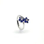 "An elegant 18ct white gold ring with a crossover shank featuring two clusters of five round bright blue sapphires, each cluster surrounds a single white sparkling diamond, creating a captivating and sophisticated design." Angled View.