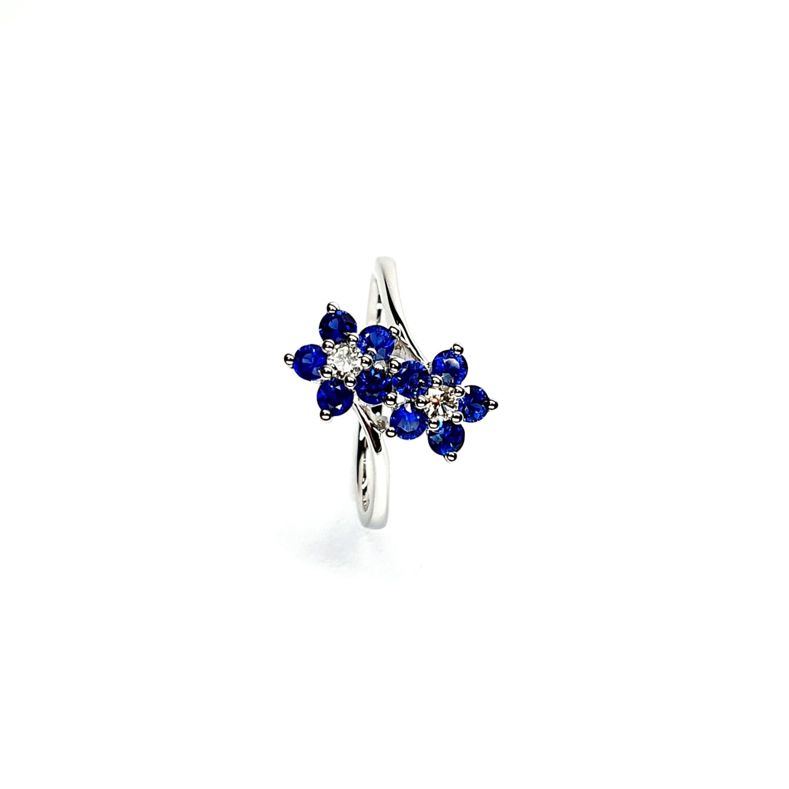 "An elegant 18ct white gold ring with a crossover shank featuring two clusters of five round bright blue sapphires, each cluster surrounds a single white sparkling diamond, creating a captivating and sophisticated design." Top View.