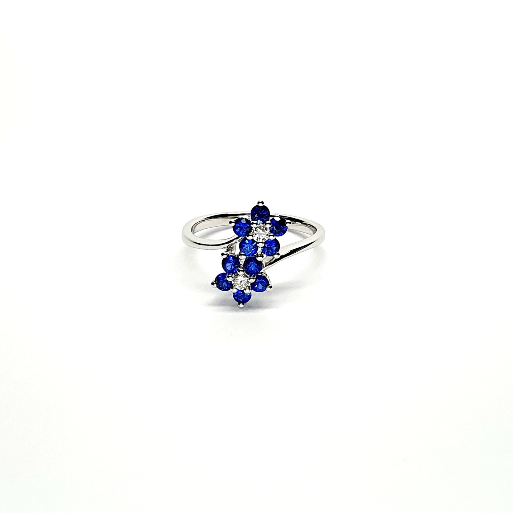 "An elegant 18ct white gold ring with a crossover shank featuring two clusters of five round bright blue sapphires, each cluster surrounds a single white sparkling diamond, creating a captivating and sophisticated design." Lying down View.