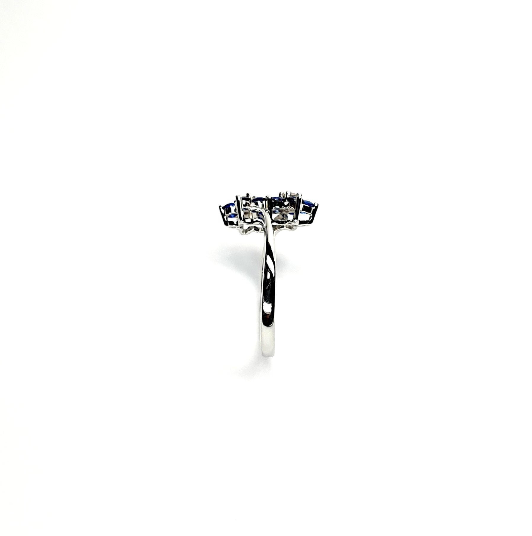 "An elegant 18ct white gold ring with a crossover shank featuring two clusters of five round bright blue sapphires, each cluster surrounds a single white sparkling diamond, creating a captivating and sophisticated design." Edge View.