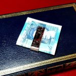 Hallmarked sterling silver scroll engraved money clip with a plain rectangular centred cartouche suitable for initials—front view pictured with a five-pound note on a blue book.