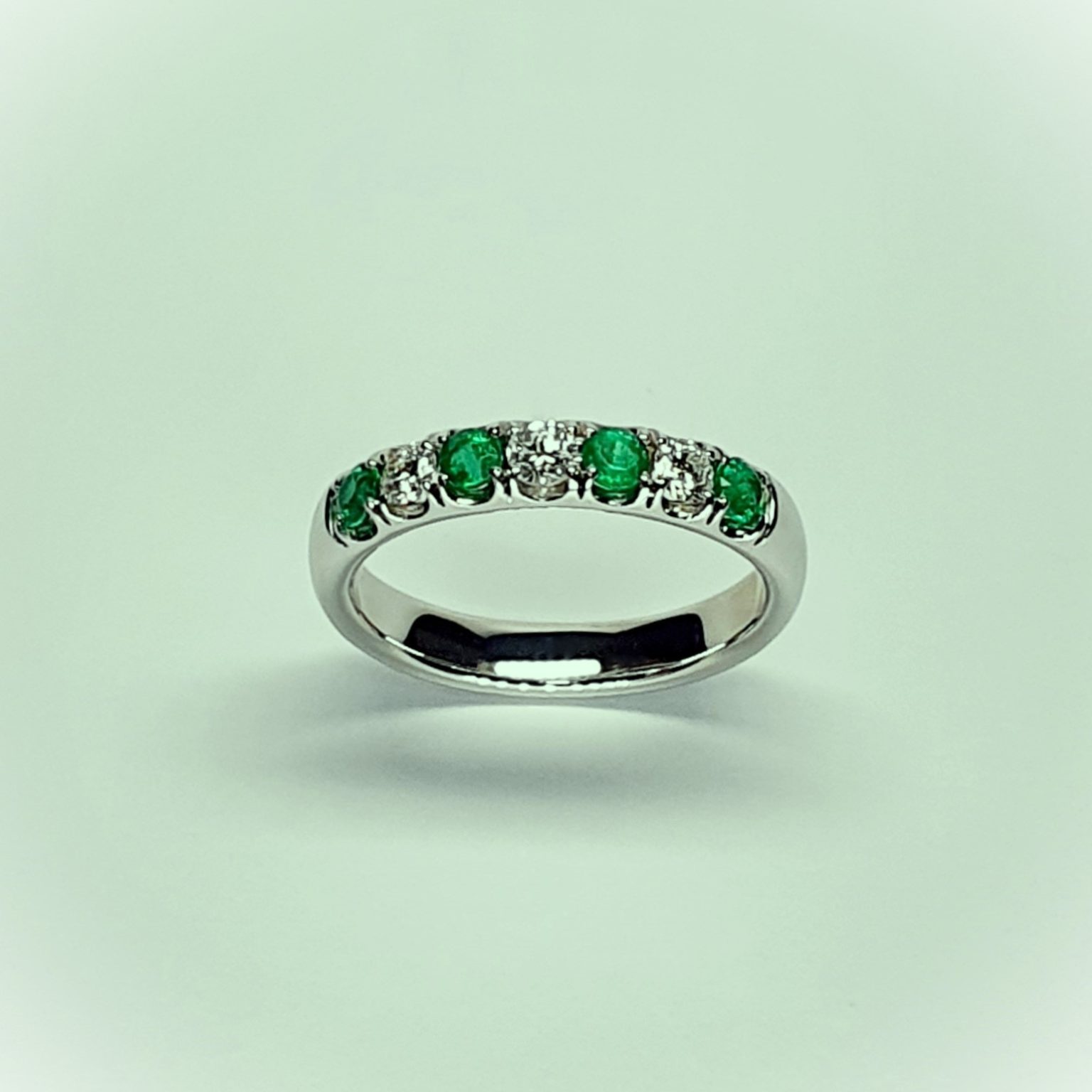 18ct White Gold Emerald & Diamond Eternity Ring RA00685 Four 2.8mm round emeralds and three 2.8mm round diamonds ring standing up to the front angled facing to the front. With a green tinted background.
