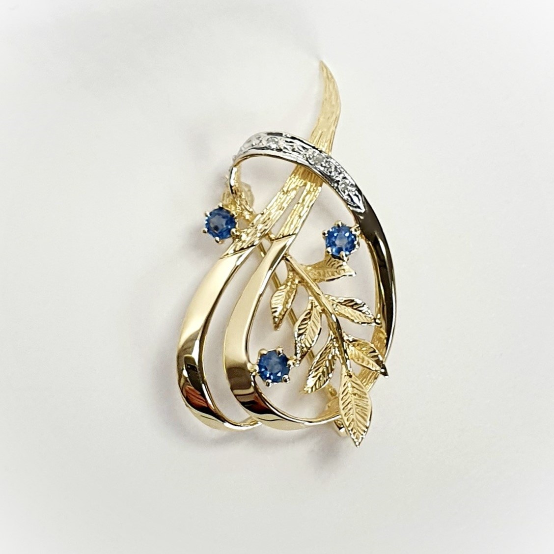 9ct yellow gold brooch with a sprig of seven leaves coming through a floral swirl of polished and barked yellow gold with three diamonds set into a small white rhodium section 