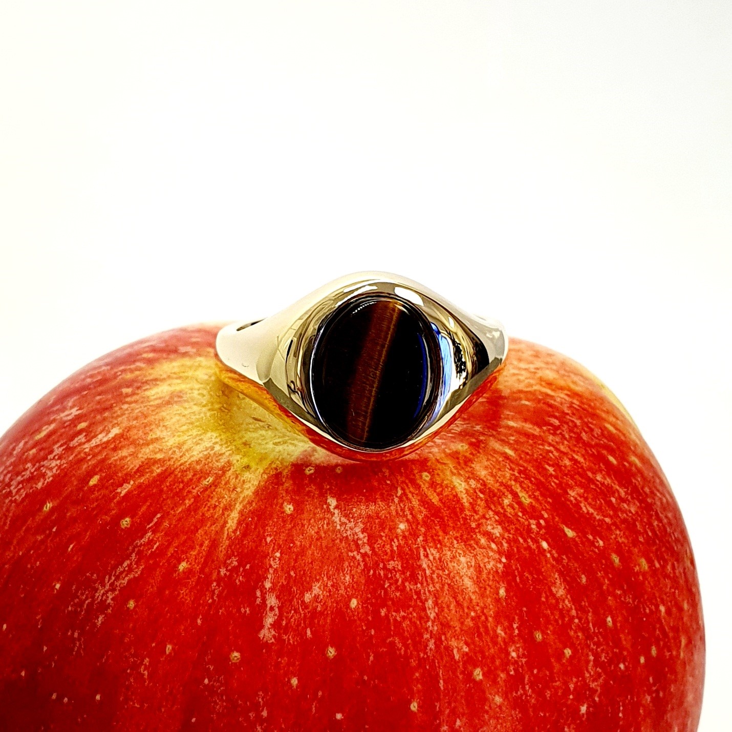 The City of London Jewellers presents the Eyemouth heavy 9ct yellow gold 12mm x 10mm oval Tigers Eye with a slightly curved top rub-over set into a plain polished signet ring, angled view laying down from the front sitting on an apple.