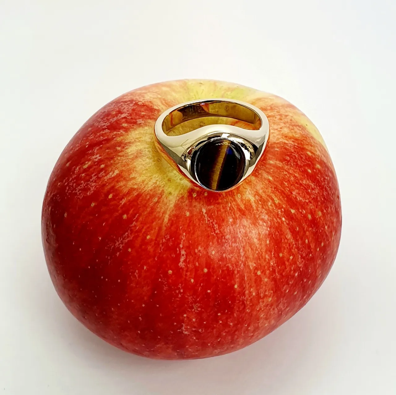 The City of London Jewellers presents the Eyemouth heavy 9ct yellow gold 12mm x 10mm oval Tigers Eye with a slightly curved top rub-over set into a plain polished signet ring, angled view laying down from the front sitting on an apple slight angle from above.