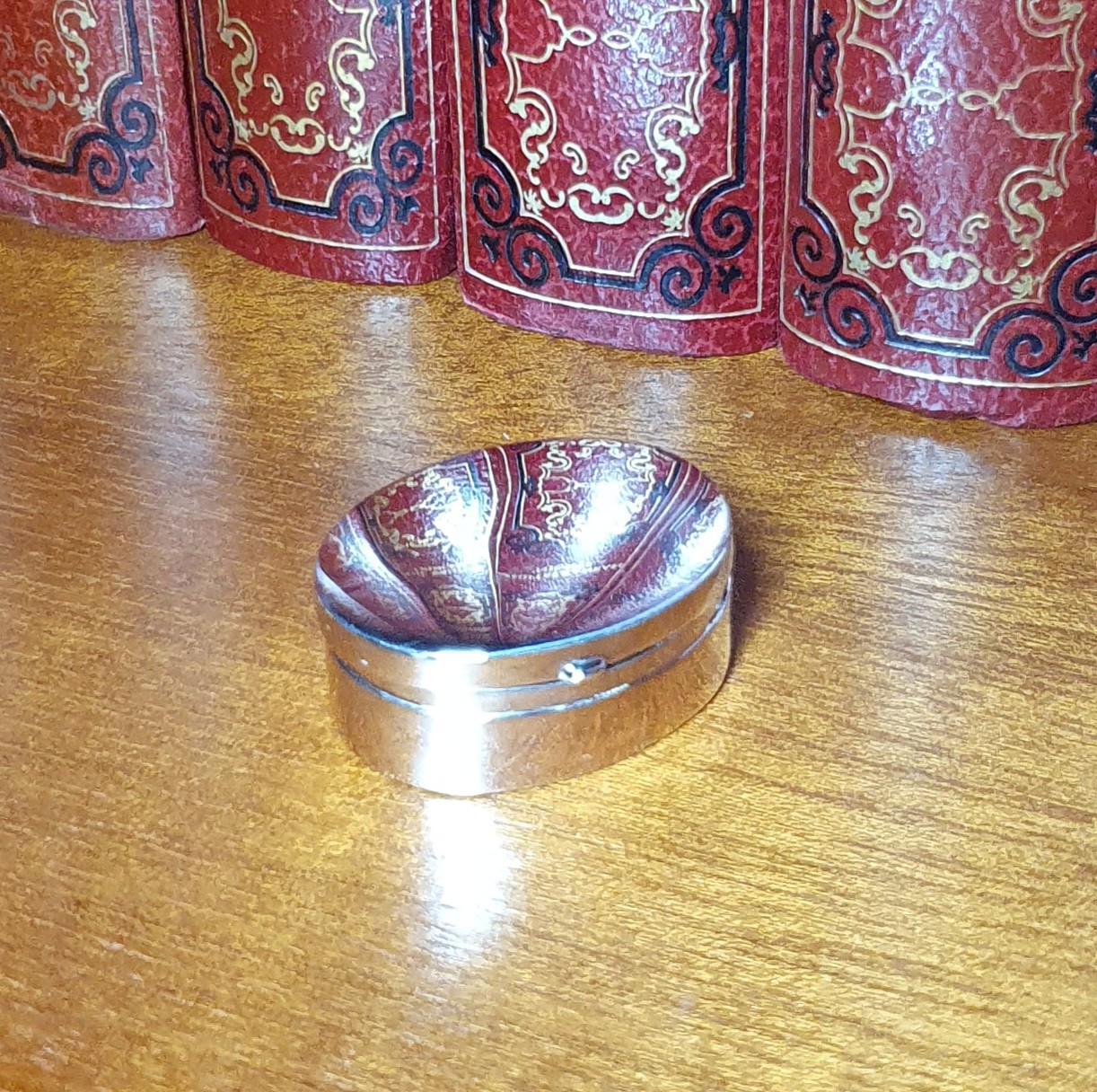 Plain polished oval genuine silver pill/trinket box pictured closed sitting on book shelf.