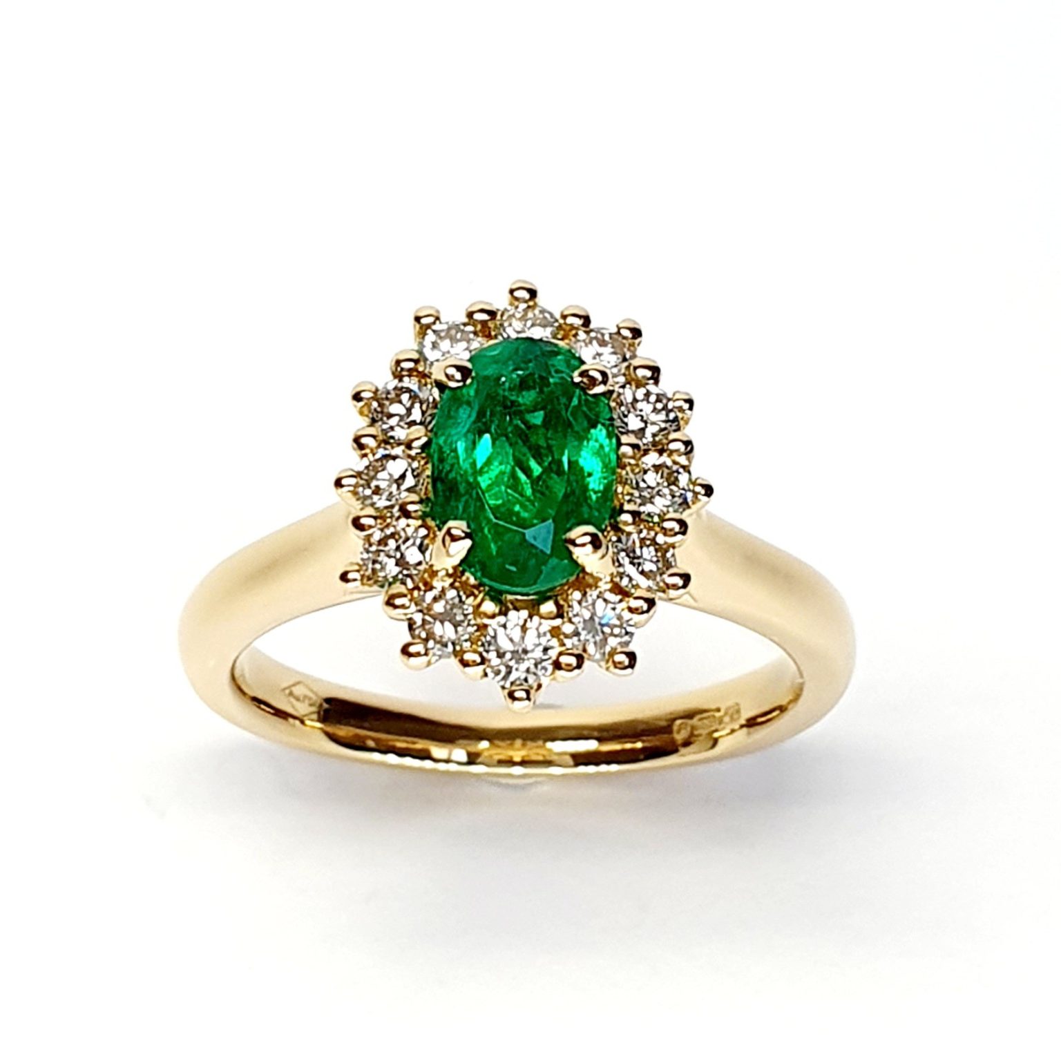 At the heart of this exquisite 18ct yellow gold ring lies a resplendent 7mm x 5mm oval emerald weighing an impressive 0.84 carats and exuding an enchanting green hue. Its captivating charm captures the essence of nature's beauty, with twelve dazzling round brilliant-cut diamonds, totalling 0.41 carats, surrounding the emerald. Each stone is meticulously selected to enhance the ring's allure with unparalleled radiance and brilliance, and the basket-wire setting measures 13mm x 10mm.RA00696.
