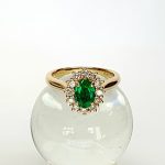 At the heart of this exquisite 18ct yellow gold ring lies a resplendent 7mm x 5mm oval emerald weighing an impressive 0.84 carats and exuding an enchanting green hue. Its captivating charm captures the essence of nature's beauty, with twelve dazzling round brilliant-cut diamonds, totalling 0.41 carats, surrounding the emerald. Each stone is meticulously selected to enhance the ring's allure with unparalleled radiance and brilliance, and the basket-wire setting measures 13mm x 10mm.RA00696.