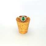 At the heart of this exquisite 18ct yellow gold ring lies a resplendent 7mm x 5mm oval emerald weighing an impressive 0.84 carats and exuding an enchanting green hue. Its captivating charm captures the essence of nature's beauty, with twelve dazzling round brilliant-cut diamonds, totalling 0.41 carats, surrounding the emerald. Each stone is meticulously selected to enhance the ring's allure with unparalleled radiance and brilliance, and the basket-wire setting measures 13mm x 10mm.RA00696.