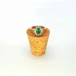 At the heart of this exquisite 18ct yellow gold ring lies a resplendent 7mm x 5mm oval emerald weighing an impressive 0.84 carats and exuding an enchanting green hue. Its captivating charm captures the essence of nature's beauty, with twelve dazzling round brilliant-cut diamonds, totalling 0.41 carats, surrounding the emerald. Each stone is meticulously selected to enhance the ring's allure with unparalleled radiance and brilliance, and the basket-wire setting measures 13mm x 10mm.RA00696.