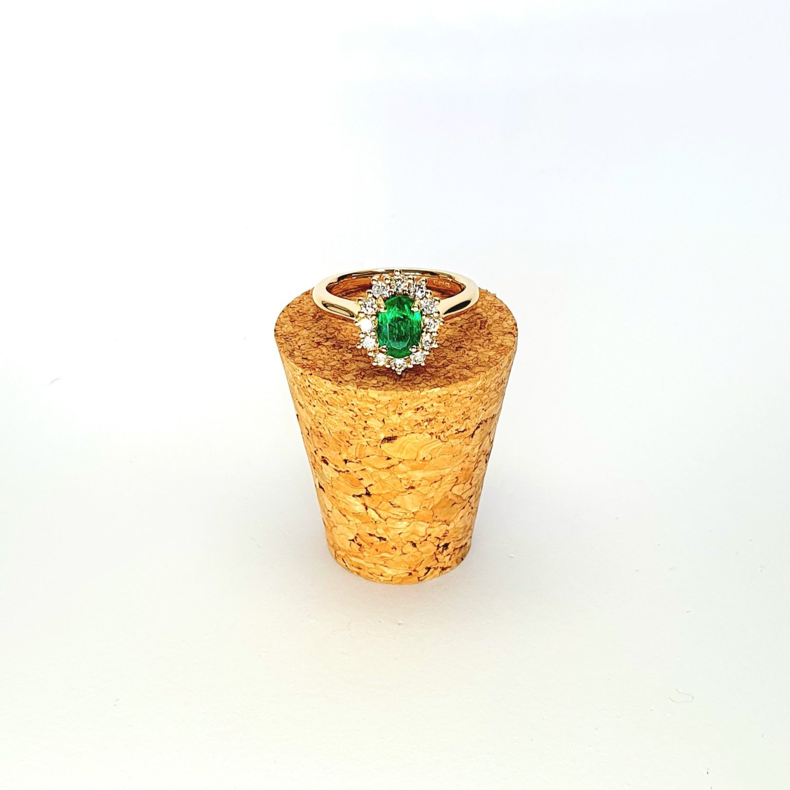 At the heart of this exquisite 18ct yellow gold ring lies a resplendent 7mm x 5mm oval emerald weighing an impressive 0.84 carats and exuding an enchanting green hue. Its captivating charm captures the essence of nature's beauty, with twelve dazzling round brilliant-cut diamonds, totalling 0.41 carats, surrounding the emerald. Each stone is meticulously selected to enhance the ring's allure with unparalleled radiance and brilliance, and the basket-wire setting measures 13mm x 10mm.RA00696.