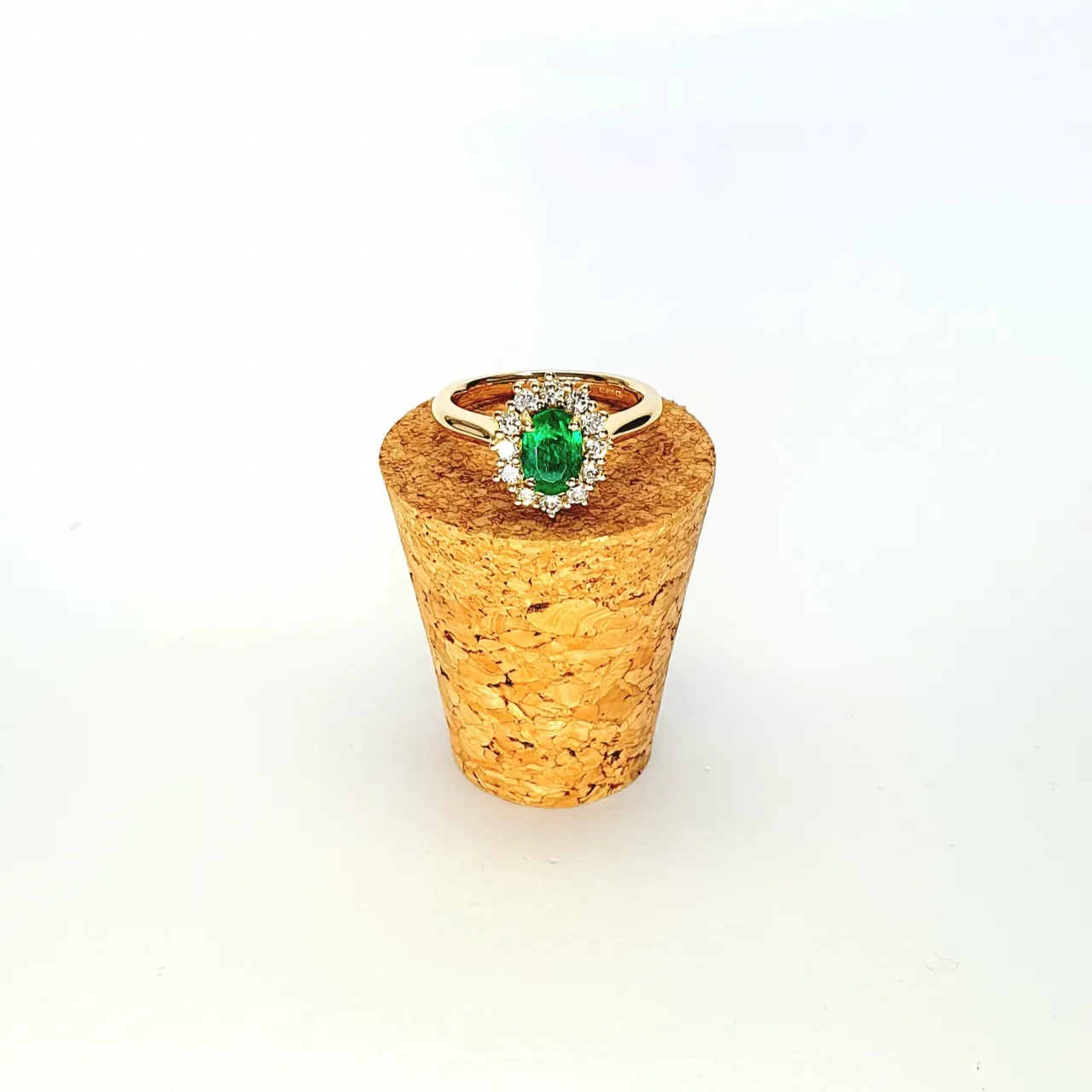 At the heart of this exquisite 18ct yellow gold ring lies a resplendent 7mm x 5mm oval emerald weighing an impressive 0.84 carats and exuding an enchanting green hue. Its captivating charm captures the essence of nature's beauty, with twelve dazzling round brilliant-cut diamonds, totalling 0.41 carats, surrounding the emerald. Each stone is meticulously selected to enhance the ring's allure with unparalleled radiance and brilliance, and the basket-wire setting measures 13mm x 10mm.RA00696.