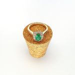 At the heart of this exquisite 18ct yellow gold ring lies a resplendent 7mm x 5mm oval emerald weighing an impressive 0.84 carats and exuding an enchanting green hue. Its captivating charm captures the essence of nature's beauty, with twelve dazzling round brilliant-cut diamonds, totalling 0.41 carats, surrounding the emerald. Each stone is meticulously selected to enhance the ring's allure with unparalleled radiance and brilliance, and the basket-wire setting measures 13mm x 10mm.RA00696.