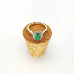 At the heart of this exquisite 18ct yellow gold ring lies a resplendent 7mm x 5mm oval emerald weighing an impressive 0.84 carats and exuding an enchanting green hue. Its captivating charm captures the essence of nature's beauty, with twelve dazzling round brilliant-cut diamonds, totalling 0.41 carats, surrounding the emerald. Each stone is meticulously selected to enhance the ring's allure with unparalleled radiance and brilliance, and the basket-wire setting measures 13mm x 10mm.RA00696.