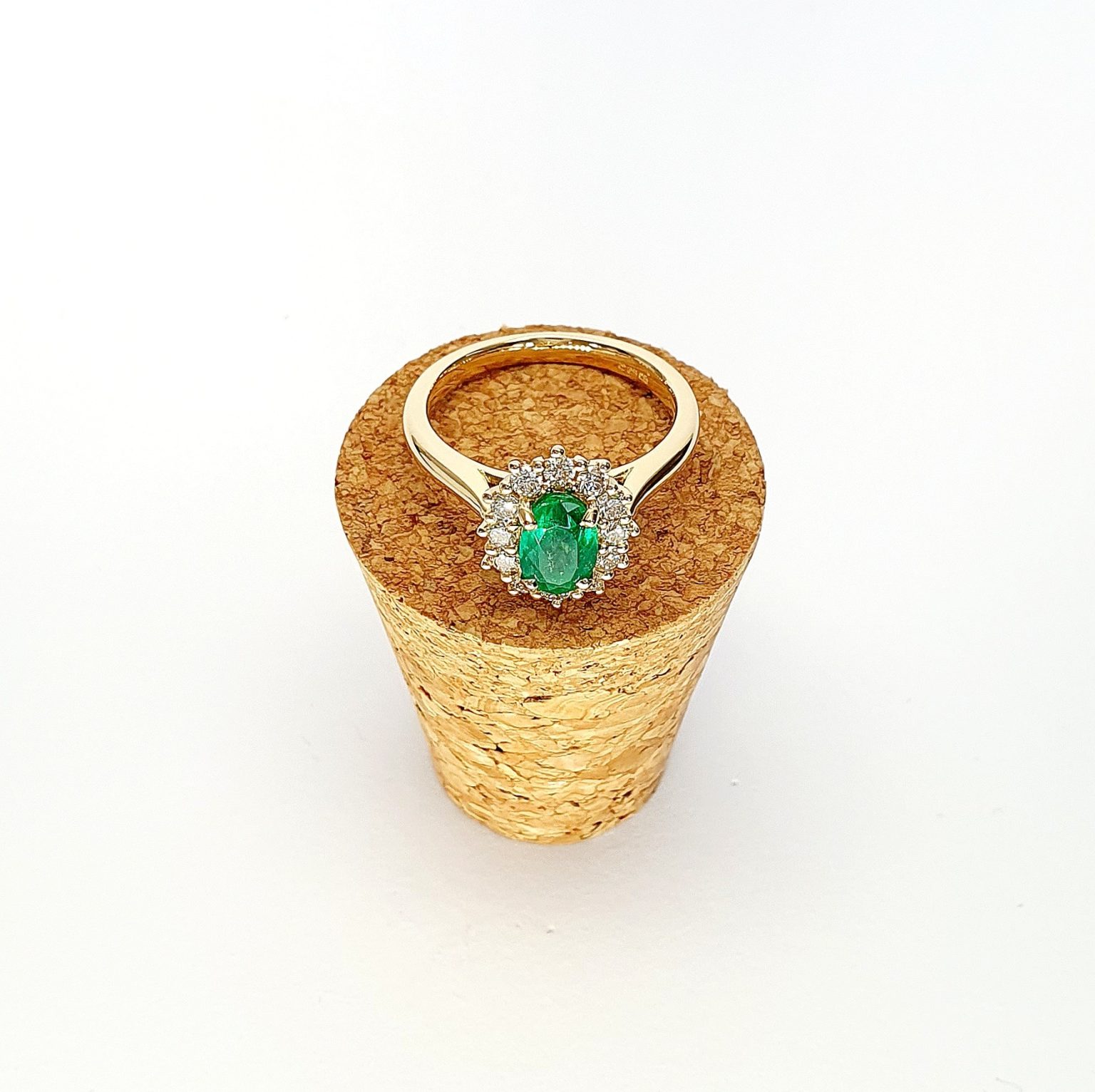 At the heart of this exquisite 18ct yellow gold ring lies a resplendent 7mm x 5mm oval emerald weighing an impressive 0.84 carats and exuding an enchanting green hue. Its captivating charm captures the essence of nature's beauty, with twelve dazzling round brilliant-cut diamonds, totalling 0.41 carats, surrounding the emerald. Each stone is meticulously selected to enhance the ring's allure with unparalleled radiance and brilliance, and the basket-wire setting measures 13mm x 10mm.RA00696.