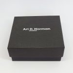 Black cardboard two part box with a black draw string pouch inside. closed.