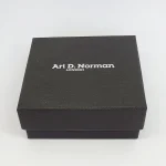Black cardboard two part box with a black draw string pouch inside. closed.