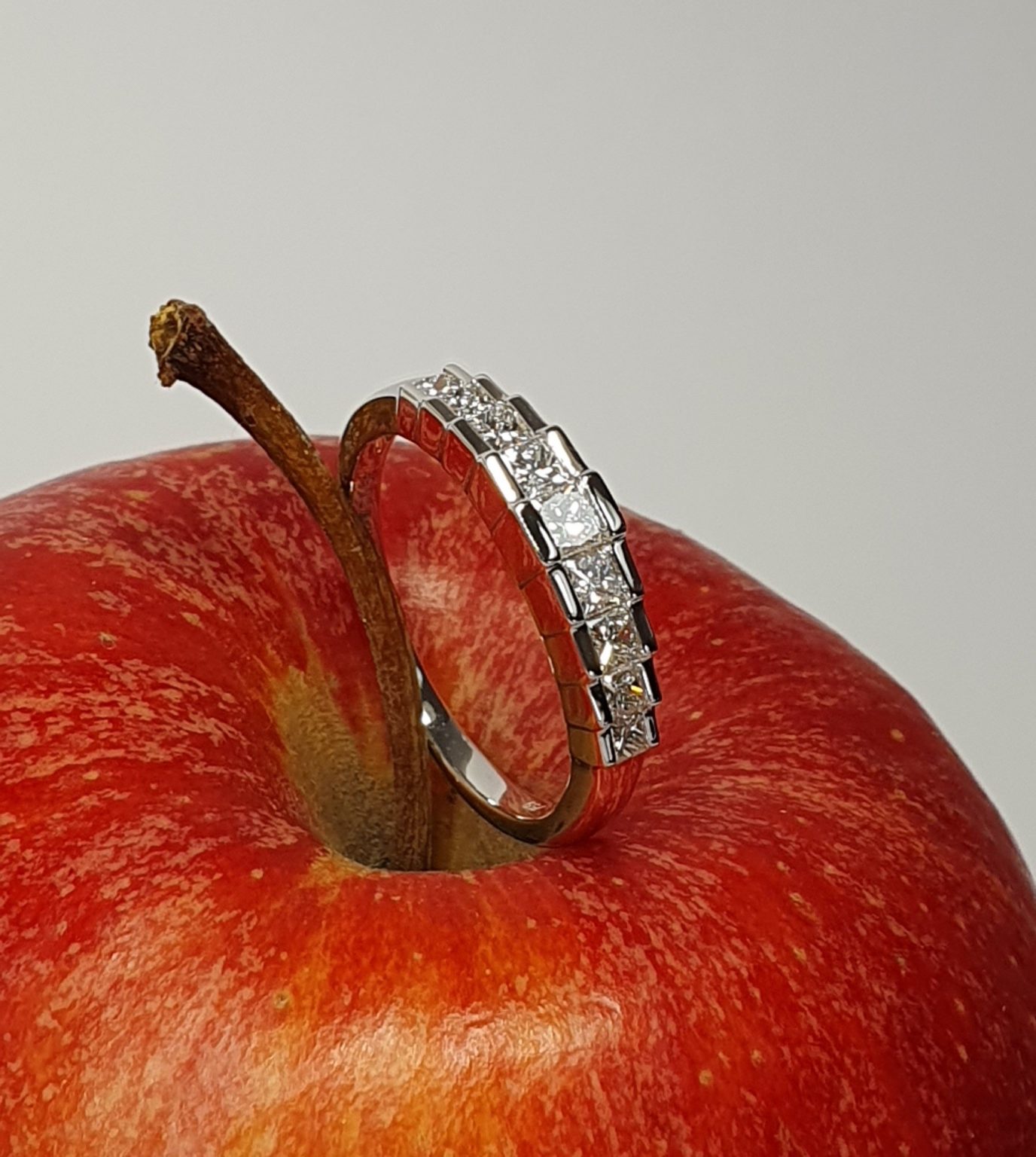 Apple Charm: Resting against the vibrant red of an apple, the ring's step design and dazzling nine graduating princess cut diamonds stones shine beautifully, symbolizing elegance and sophistication.