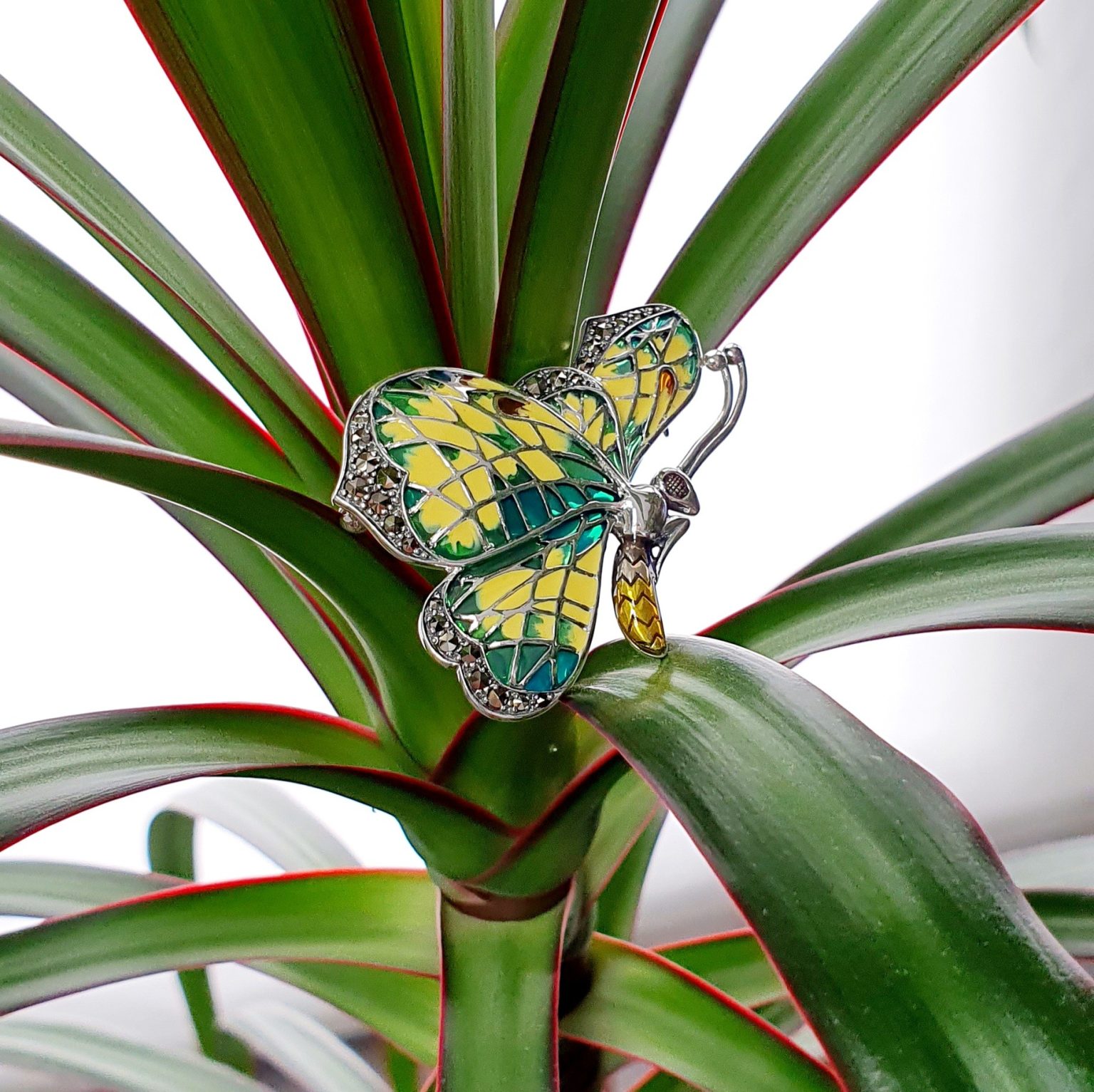 Lemon Butterfly Brooch Sterling Silver BRS00107 Genuine 925 sterling silver butterfly brooch with lemon and pale green colour Plique-à-jour enamel wings with real marcasite set to the tips, the body and eye with a golden yellow enamel with two long polished silver antennae. Pictured sitting on a plant.
