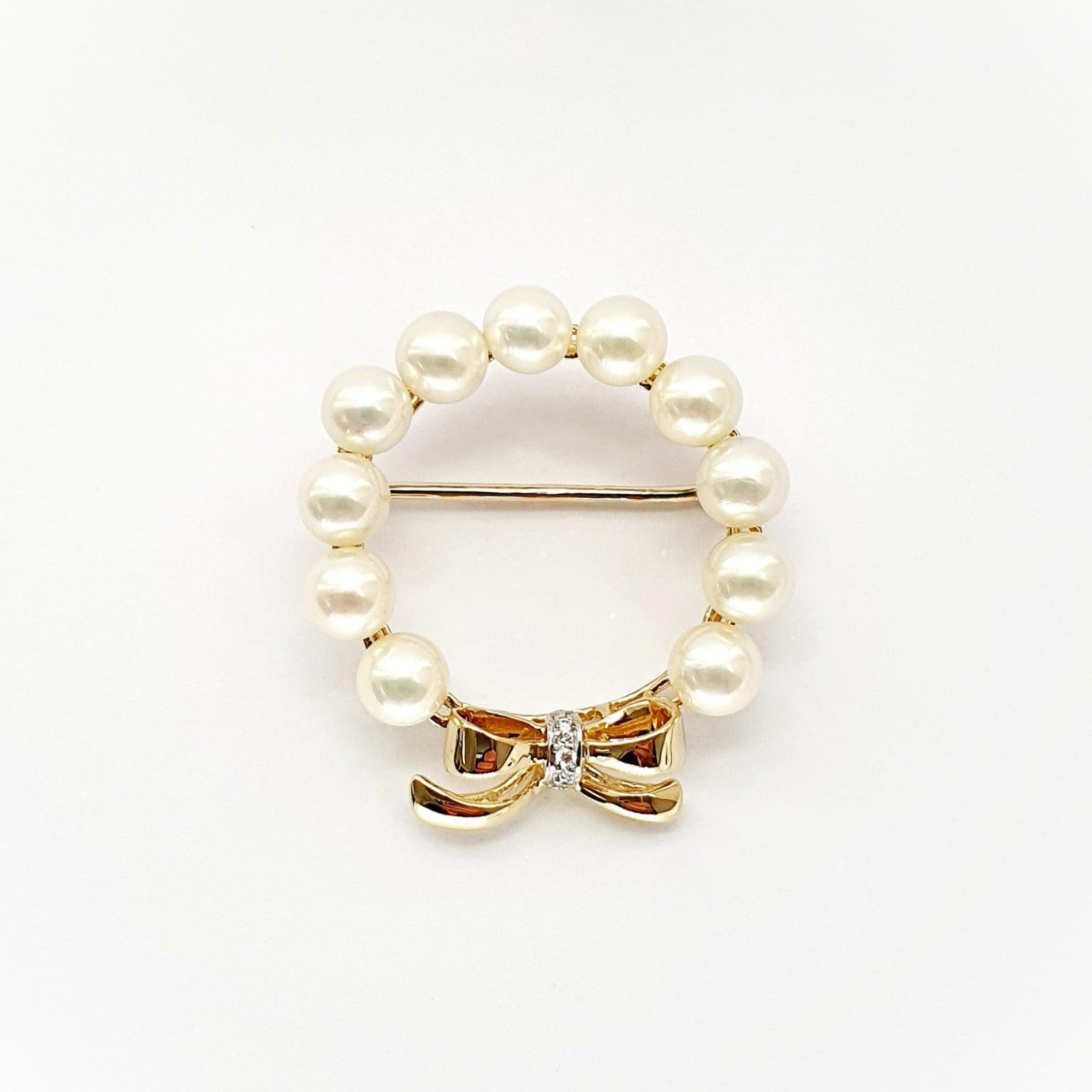 A 9ct yellow gold brooch with a circle of eleven 5mm round pearls leading to a bow to the bottom with three petite round diamonds set to the centre of the bow. Front view showing the pin threw the centre.