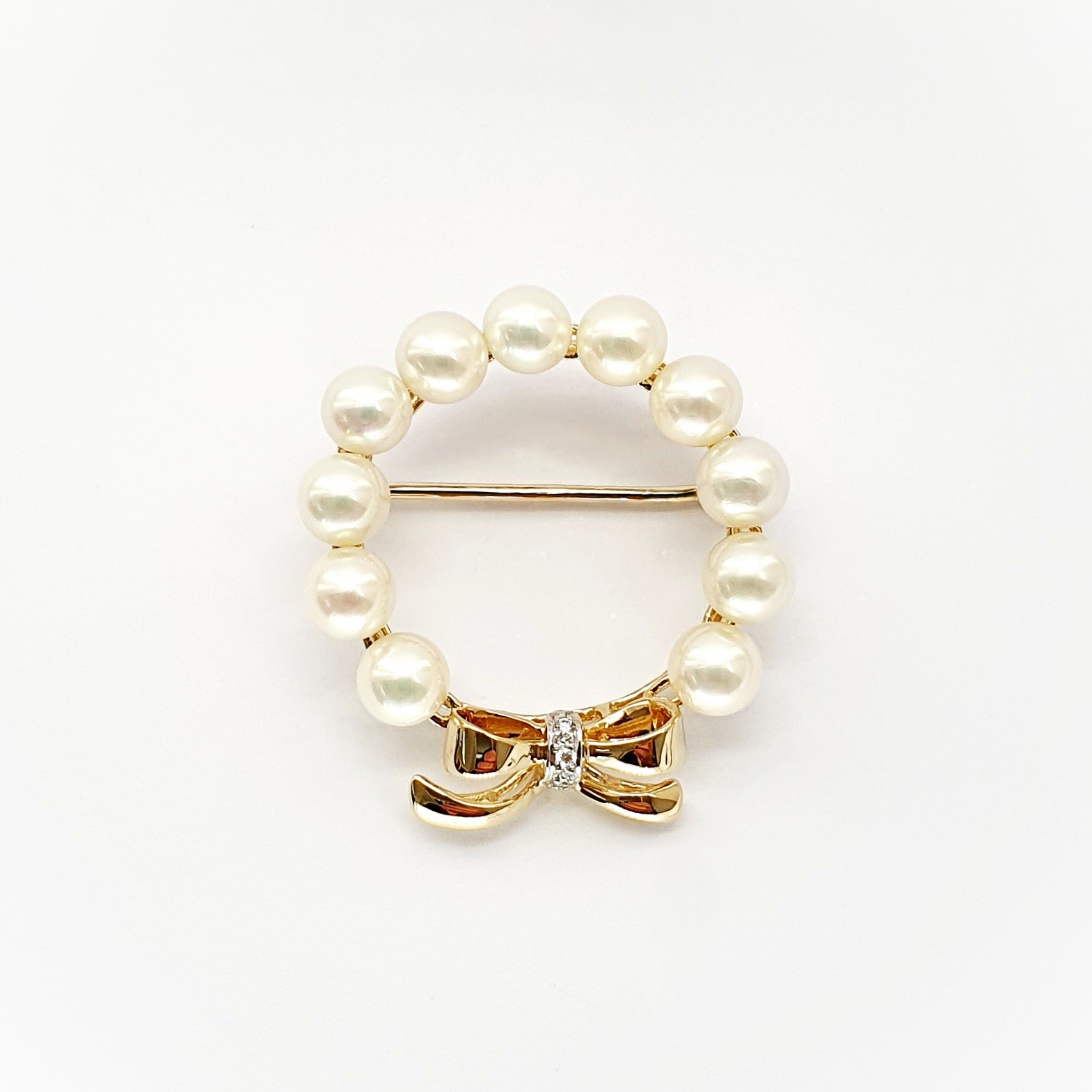 A 9ct yellow gold brooch with a circle of eleven 5mm round pearls leading to a bow to the bottom with three petite round diamonds set to the centre of the bow. Front view showing the pin threw the centre.