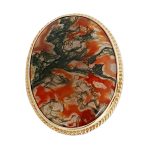 This stunning brooch features a domed oval moss agate stone semi-translucent, exhibits captivating dendritic inclusions. As a result, the stone displays mesmerizing swirls of orangish-red and deep green., exquisitely set in a 9ct yellow gold rub-over setting it is framed by twisted gold wire and includes an open-back bezel to maximizes light exposure. Front view close up.