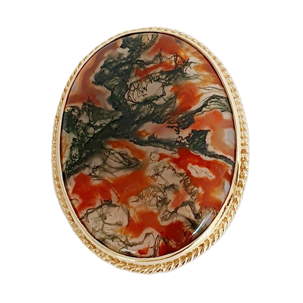 This stunning brooch features a domed oval moss agate stone semi-translucent, exhibits captivating dendritic inclusions. As a result, the stone displays mesmerizing swirls of orangish-red and deep green., exquisitely set in a 9ct yellow gold rub-over setting it is framed by twisted gold wire and includes an open-back bezel to maximizes light exposure. Front view close up.