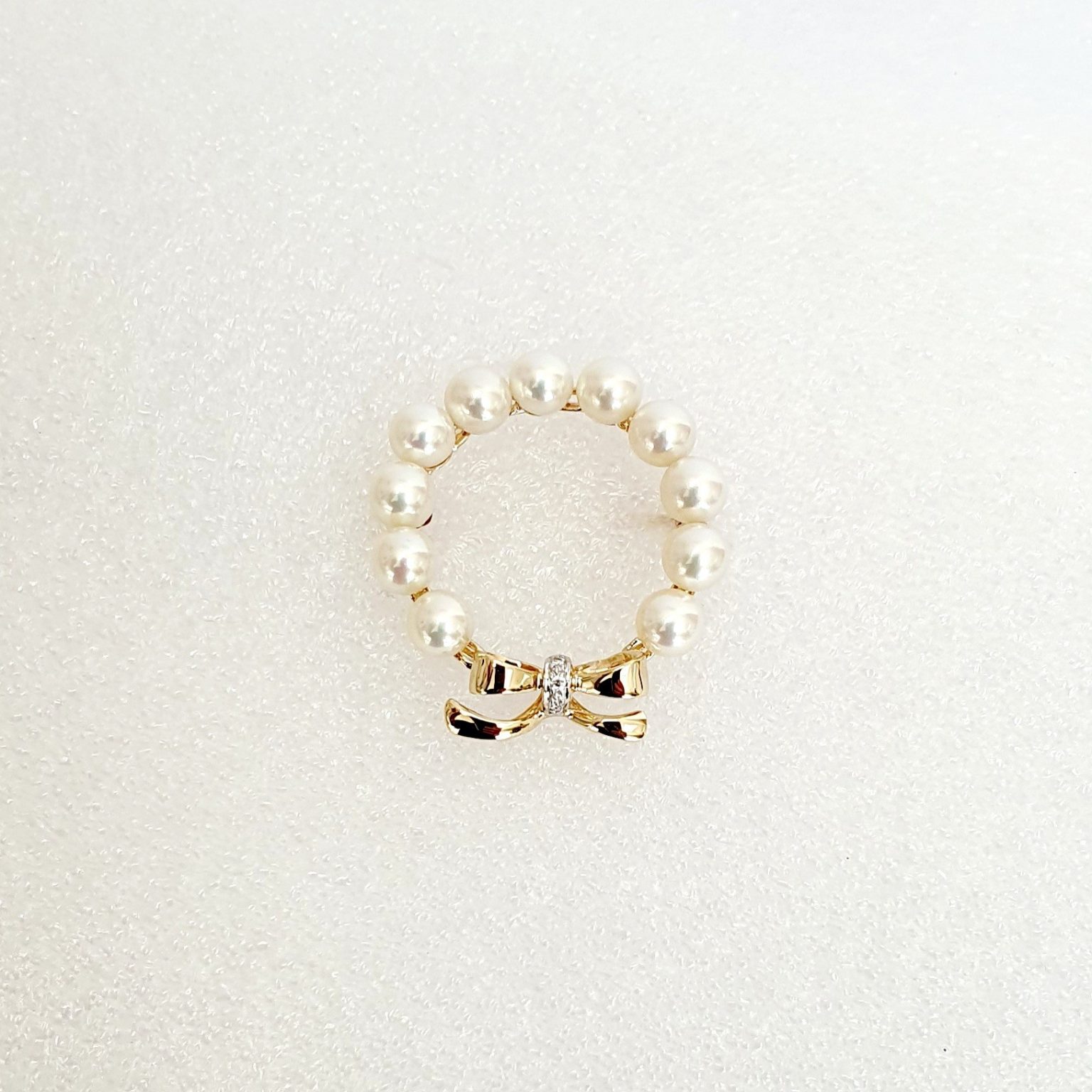 A 9ct yellow gold brooch with a circle of eleven 5mm round pearls leading to a bow to the bottom with three petite round diamonds set to the centre of the bow. showing pinned to a white background.