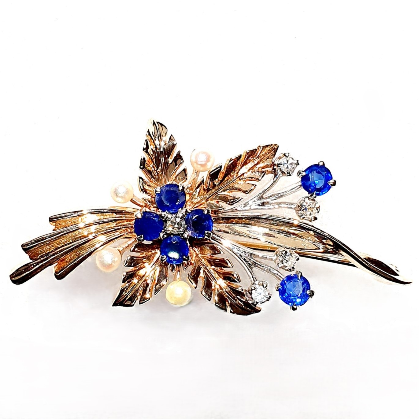 9ct Yellow Gold Ruby-Pearl Flower Brooch (36mm x 24mm) - Captivating floral elegance in 9ct yellow & white gold adorned with blue sapphires and cultured pearls. A masterpiece by The City of London Jewellers, showcasing nature's beauty and craftsmanship. One centre four-fern style leaf design with four sapphires and a diamond in the centre with a total of ten stems crossing behind set with two bright blue sapphires and a stem of a further four diamonds and four pearls. Front view close up.