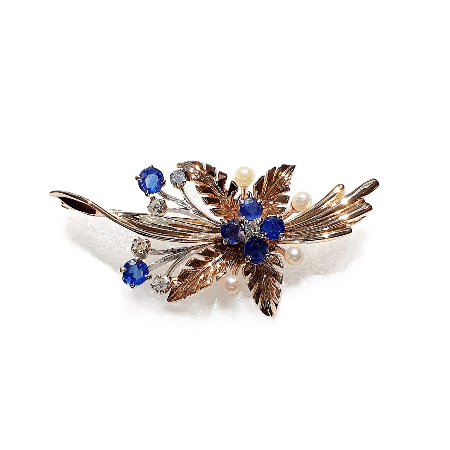 9ct Yellow Gold Ruby-Pearl Flower Brooch (36mm x 24mm) - Captivating floral elegance in 9ct yellow & white gold adorned with blue sapphires and cultured pearls. A masterpiece by The City of London Jewellers, showcasing nature's beauty and craftsmanship. One centre four-fern style leaf design with four sapphires and a diamond in the centre with a total of ten stems crossing behind set with two bright blue sapphires and a stem of a further four diamonds and four pearls. Front view white background.