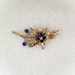 9ct Yellow Gold Ruby-Pearl Flower Brooch (36mm x 24mm) - Captivating floral elegance in 9ct yellow & white gold adorned with blue sapphires and cultured pearls. A masterpiece by The City of London Jewellers, showcasing nature's beauty and craftsmanship. One centre four-fern style leaf design with four sapphires and a diamond in the centre with a total of ten stems crossing behind set with two bright blue sapphires and a stem of a further four diamonds and four pearls. Front view matt back ground.