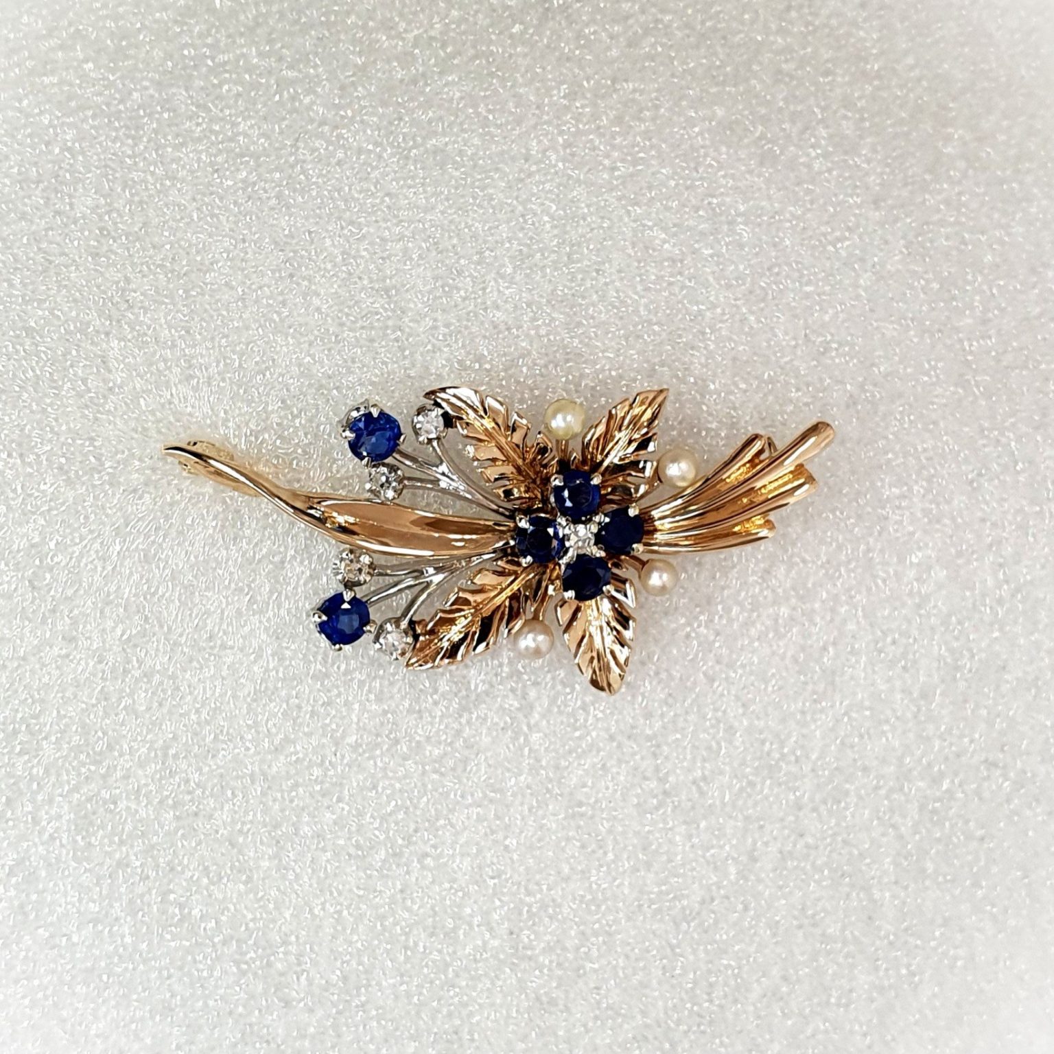 9ct Yellow Gold Ruby-Pearl Flower Brooch (36mm x 24mm) - Captivating floral elegance in 9ct yellow & white gold adorned with blue sapphires and cultured pearls. A masterpiece by The City of London Jewellers, showcasing nature's beauty and craftsmanship. One centre four-fern style leaf design with four sapphires and a diamond in the centre with a total of ten stems crossing behind set with two bright blue sapphires and a stem of a further four diamonds and four pearls. Front view matt back ground.