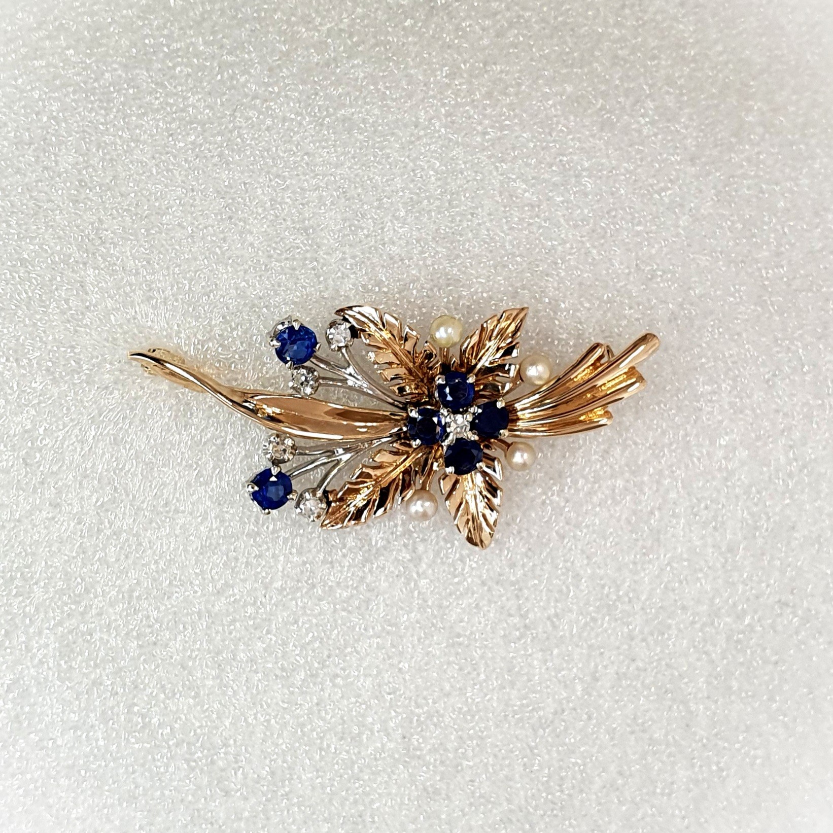 9ct Yellow Gold Ruby-Pearl Flower Brooch (36mm x 24mm) - Captivating floral elegance in 9ct yellow & white gold adorned with blue sapphires and cultured pearls. A masterpiece by The City of London Jewellers, showcasing nature's beauty and craftsmanship. One centre four-fern style leaf design with four sapphires and a diamond in the centre with a total of ten stems crossing behind set with two bright blue sapphires and a stem of a further four diamonds and four pearls. Front view matt back ground.