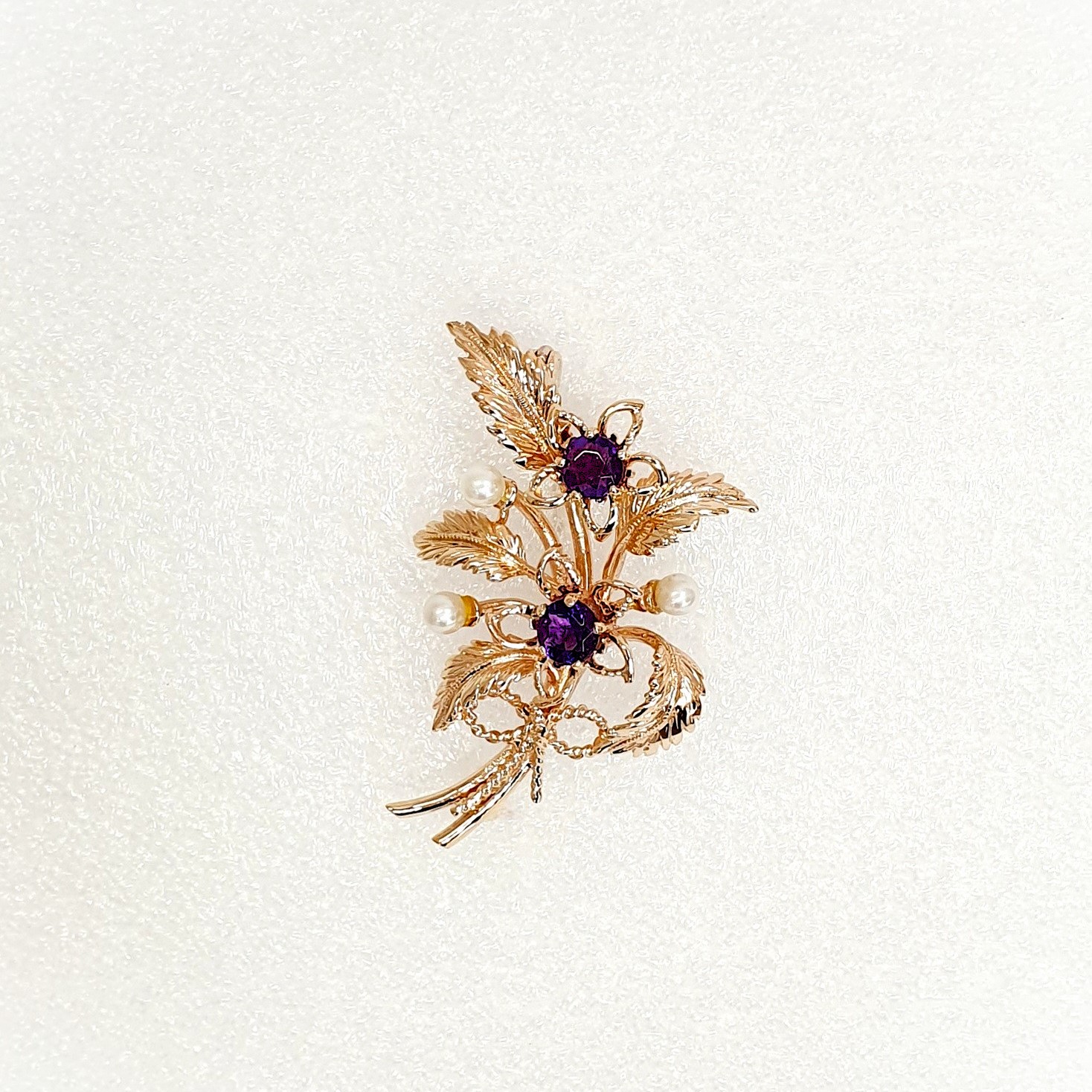 9ct rose gold flower brooch with five leaves, two round purple amethyst stones and three pearls made into a bouquet and tied to the bottom with a small twisted gold bow. Front view.