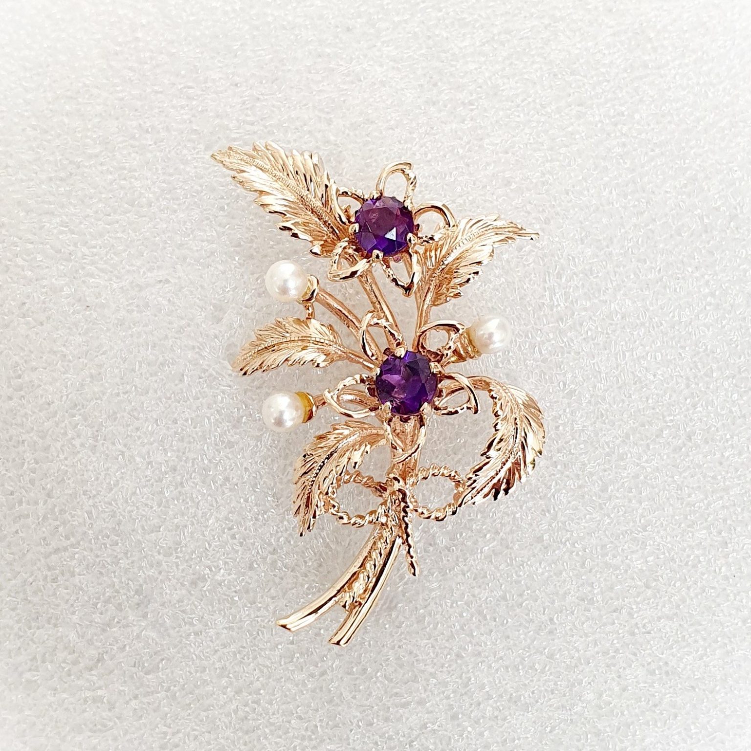 9ct rose gold flower brooch with five leaves, two round purple amethyst stones and three pearls made into a bouquet and tied to the bottom with a small twisted gold bow. Front view close up.