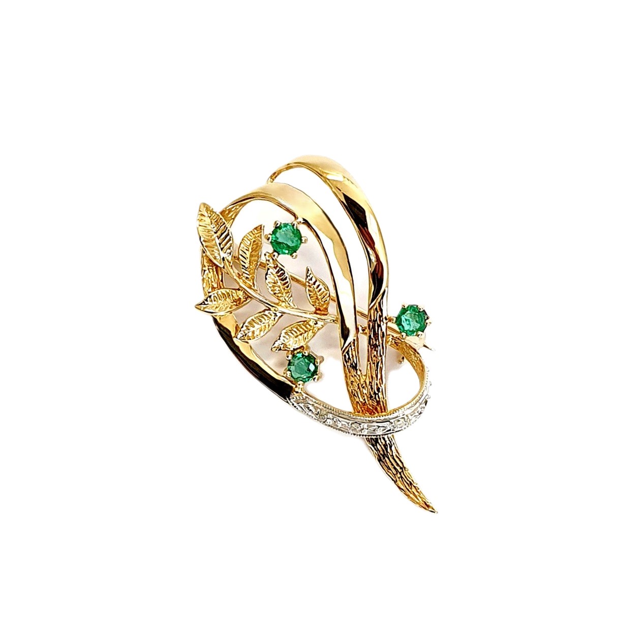 9ct yellow gold brooch with a sprig of seven leaves coming through a floral swirl of polished and barked yellow gold with three bright green emeralds & three sparkling diamonds set into a small white rhodium section, top view.