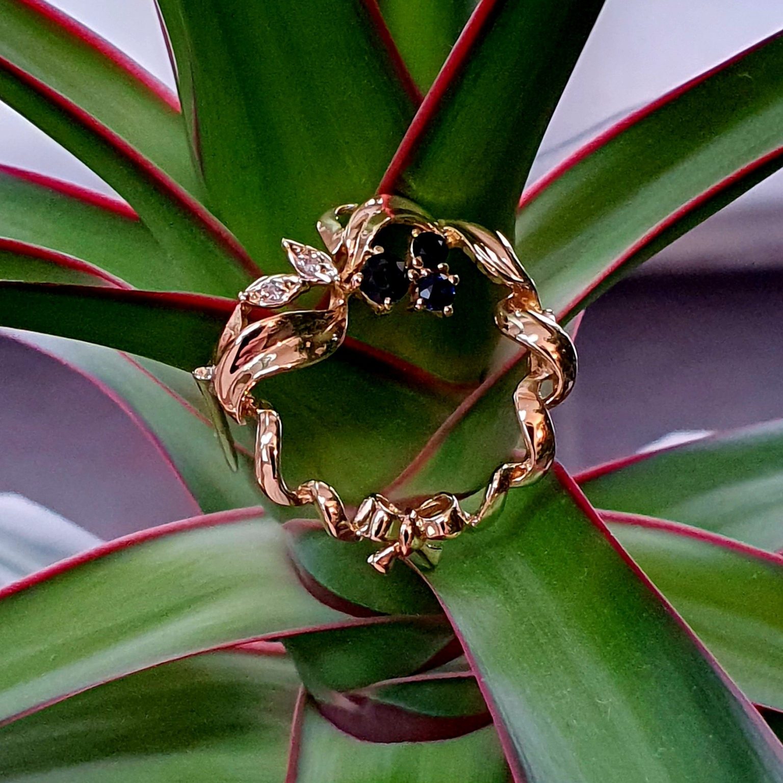 Yellow gold round ribbon with three round dark blue sapphire and two diamond leaves with a bow to the bottom centre. Pictured sitting on a plant close up.