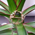 Yellow gold round ribbon with three round dark blue sapphire and two diamond leaves with a bow to the bottom centre. Pictured sitting on a plant.