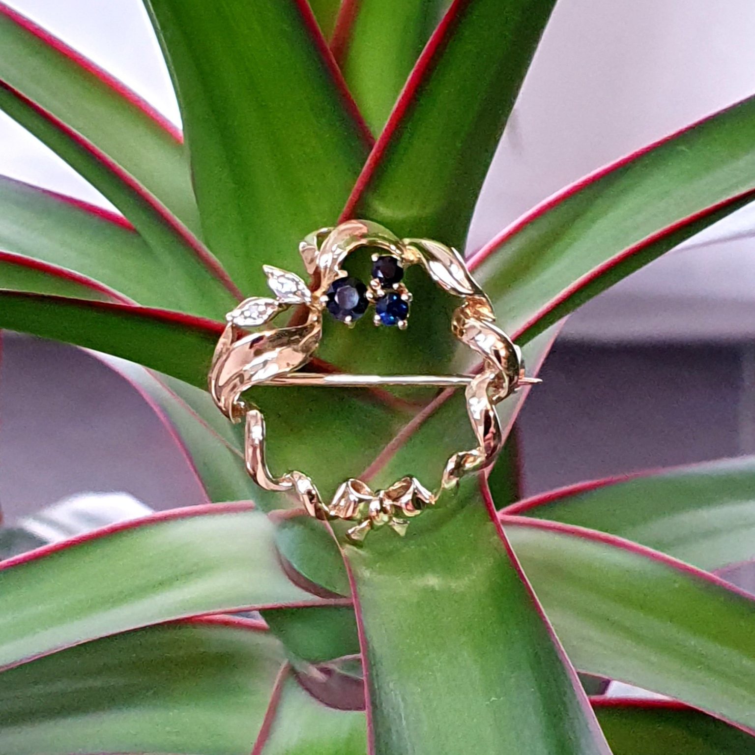 Yellow gold round ribbon with three round dark blue sapphire and two diamond leaves with a bow to the bottom centre. Pictured sitting on a plant.