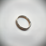 Gold wedding ring lying flat on a matte grey surface — the Sun & Snow band displayed face-up to show its rounded dual-gold design against a neutral grey background.