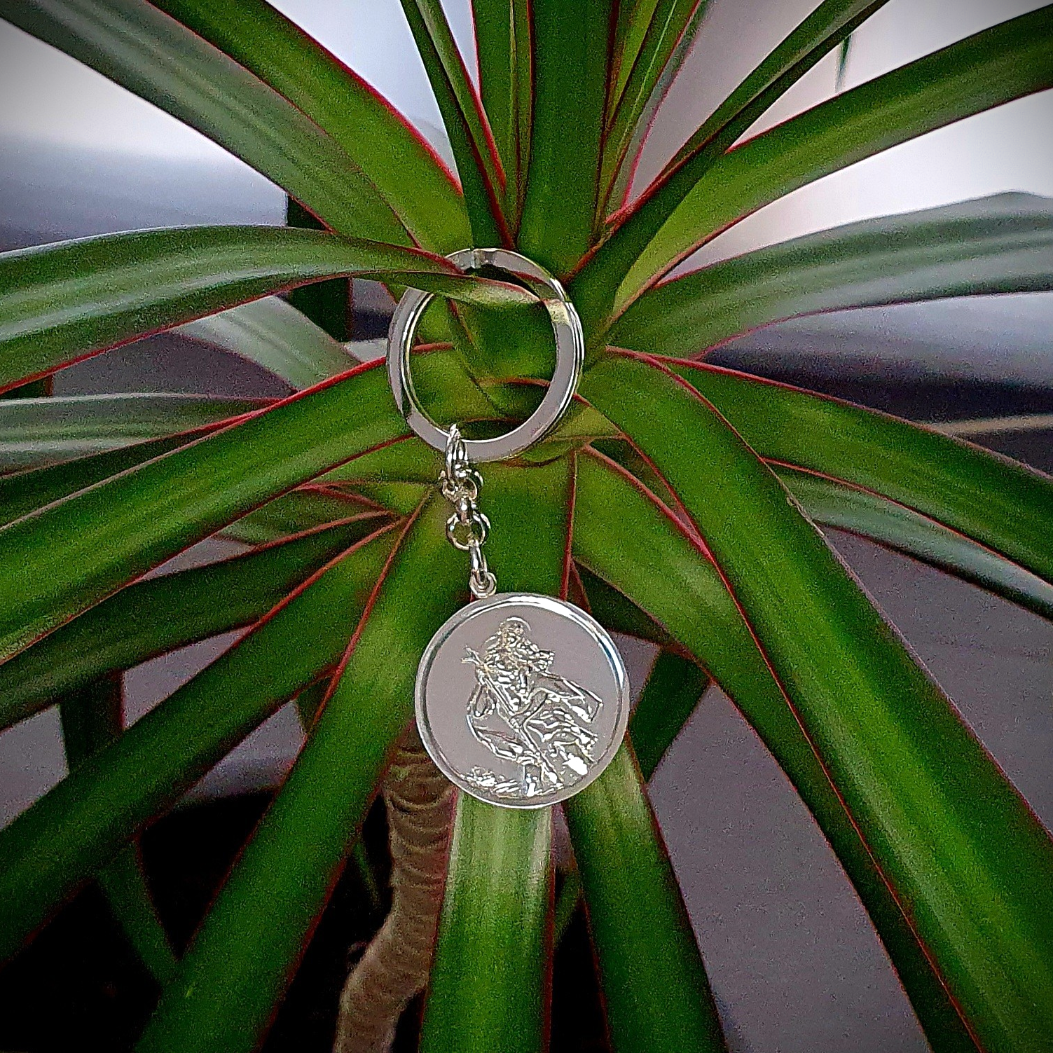 Front view on a plant: "Silver Saint Christopher keyring elegantly displayed against vibrant green leaves, highlighting its craftsmanship and design."